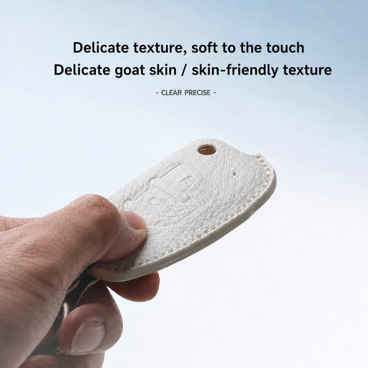 A hand holding a small white leather key fob cover with a key partially inserted, against a light blue gradient background with text describing the delicate goat skin texture.