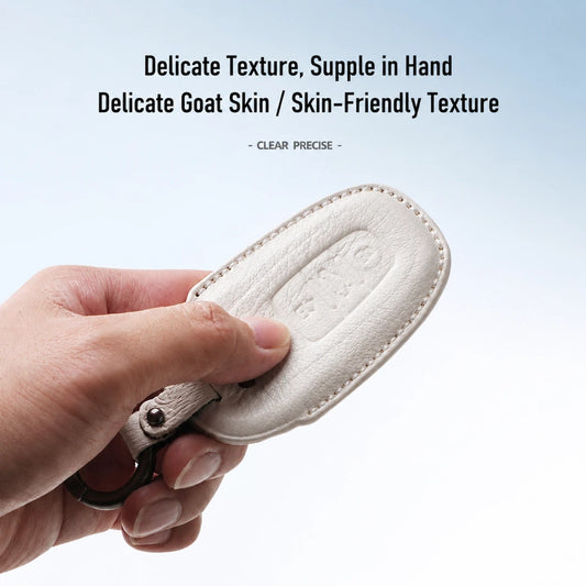 A hand holding a small oval off-white leather key fob cover or tag with visible stitching and a metal ring, against a light blue-to-white gradient background with text reading 'EXQUISITE GOAT LEATHER SKIN-LIKE TEXTURE'.