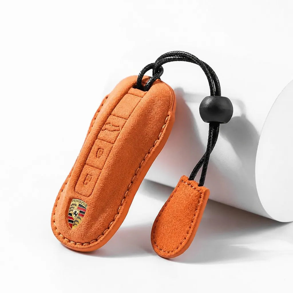 An orange suede car key fob cover with a matching keychain loop, shown on a clean white background next to a white cylindrical prop. The cover features a debossed button layout and a small gold and red Porsche crest near the bottom.