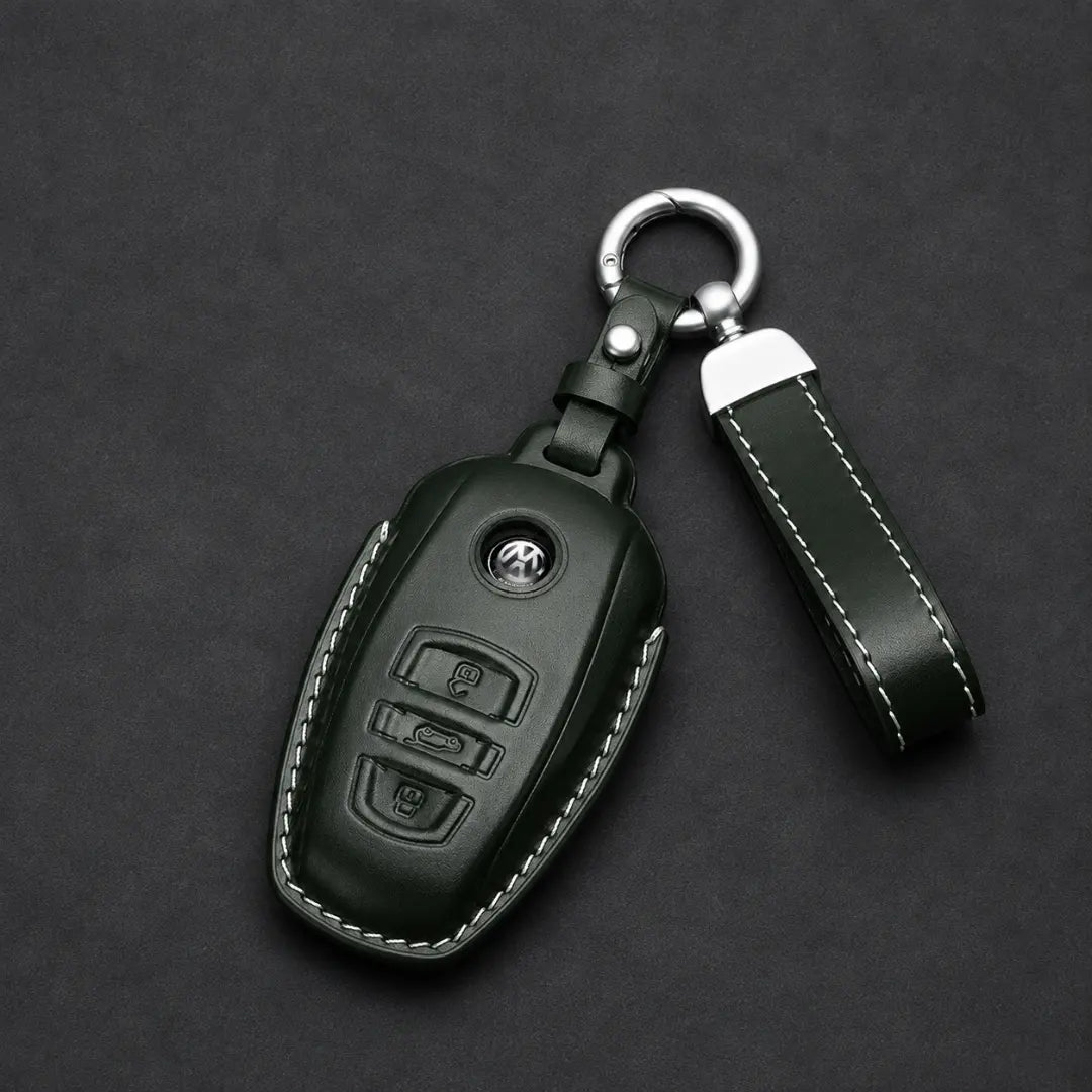 A dark green leather car key fob cover with white contrast stitching, featuring a silver metal key ring and matching leather keychain strap on a dark gray textured background.