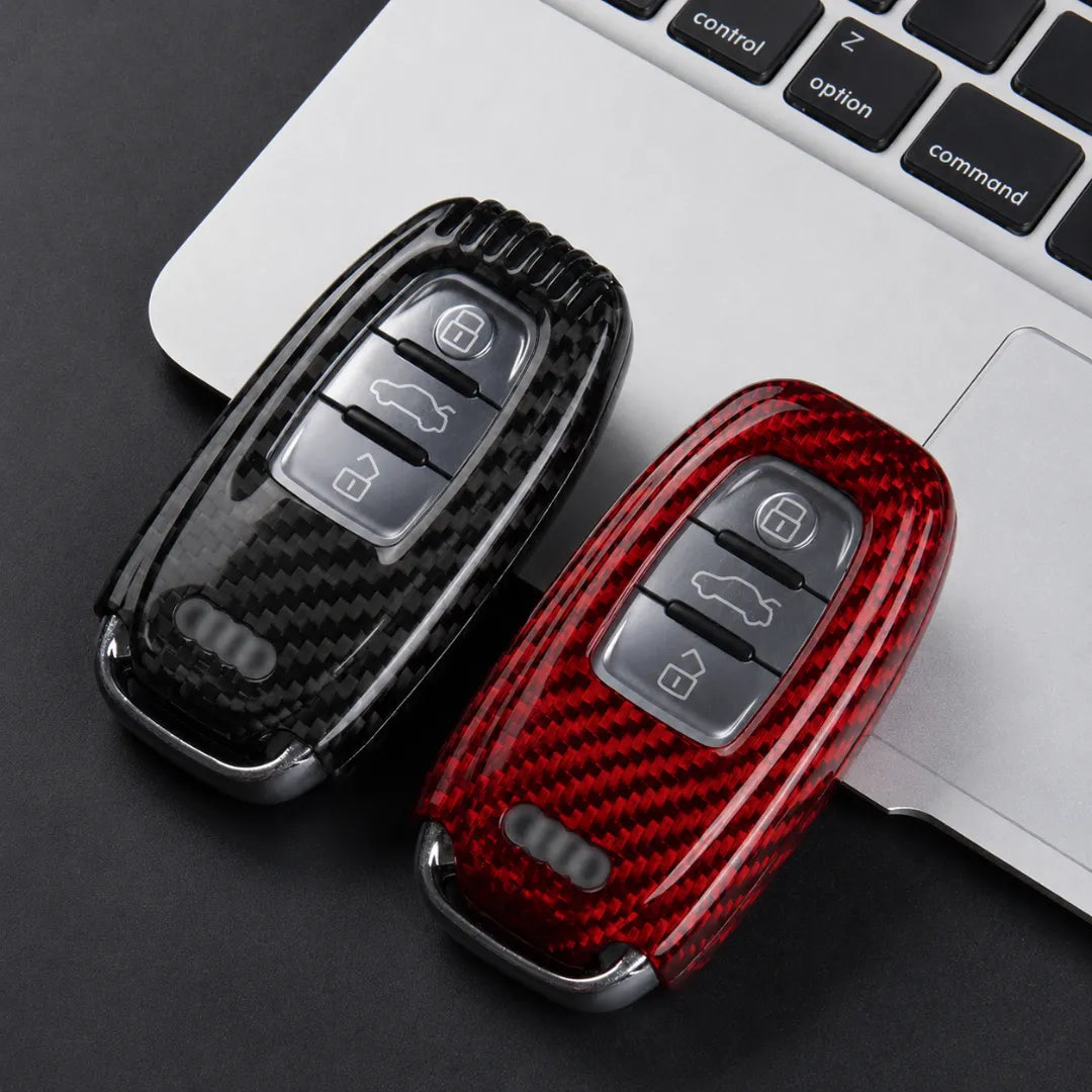 Two carbon fiber key fob covers, one black and one red, displayed on a dark surface next to a silver laptop keyboard.