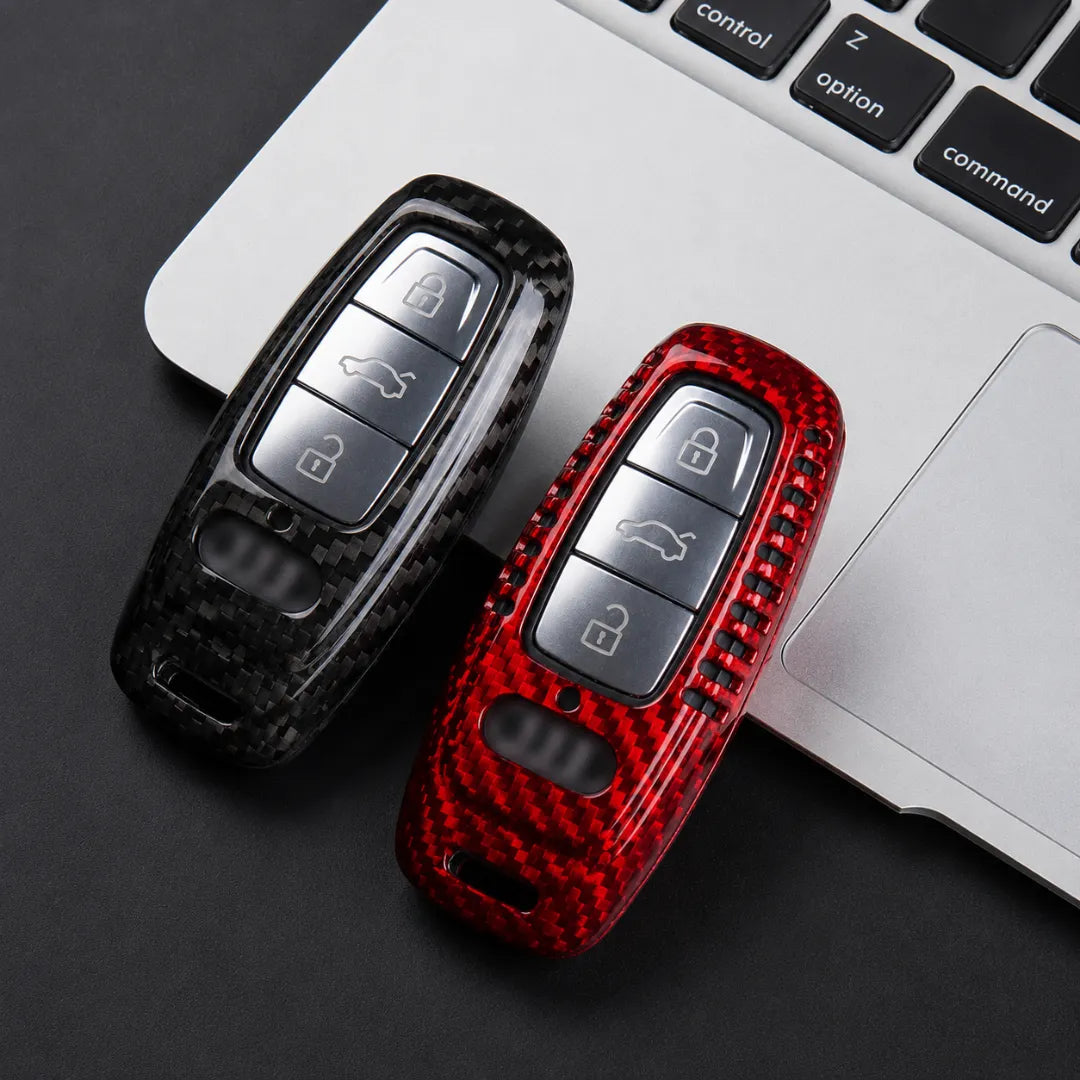 Two carbon-fiber-patterned car key fob covers in black and red, shown resting on a dark surface next to a silver laptop keyboard.