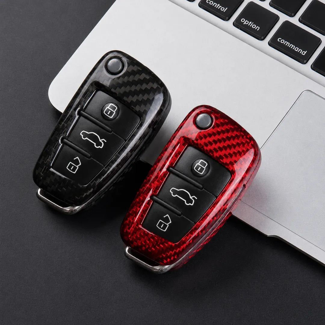 Two carbon fiber-style key fob covers, one black and one red, shown on a car key fob with a laptop keyboard in the background.