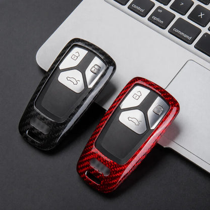 Two car key fob covers displayed on a laptop keyboard. One cover is glossy black carbon fiber textured, and the other is glossy red carbon fiber textured.