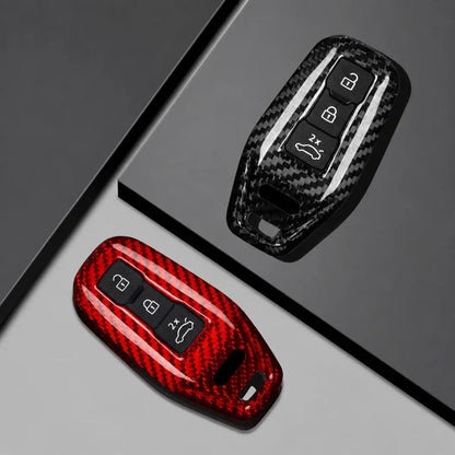Two carbon fiber-patterned car key fob covers shown on a dark gray and light gray geometric background. One cover is black with a glossy carbon fiber weave and white stripe accents, and the other is red with a matching carbon fiber weave pattern.