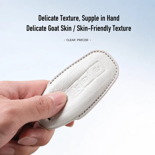 A hand holds a small white leather key fob cover against a light blue gradient background, with text above reading 'Delicate Texture, Supple in Hand' and 'Delicate Goat Skin / Skin-Friendly Texture'.