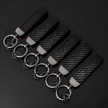 Six black leather keychains with a carbon-fiber textured pattern, silver-toned metal accents, and interlocking key rings, arranged diagonally on a dark reflective surface.