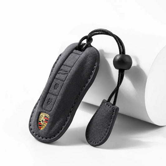 A black suede car key fob cover with a stitched border, featuring a gold and red Porsche crest emblem near the bottom. The cover is shown on a white cylindrical prop against a clean white background.