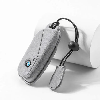 A light gray suede car key fob cover with a black adjustable cord and a small BMW logo, displayed on a white cylindrical prop.
