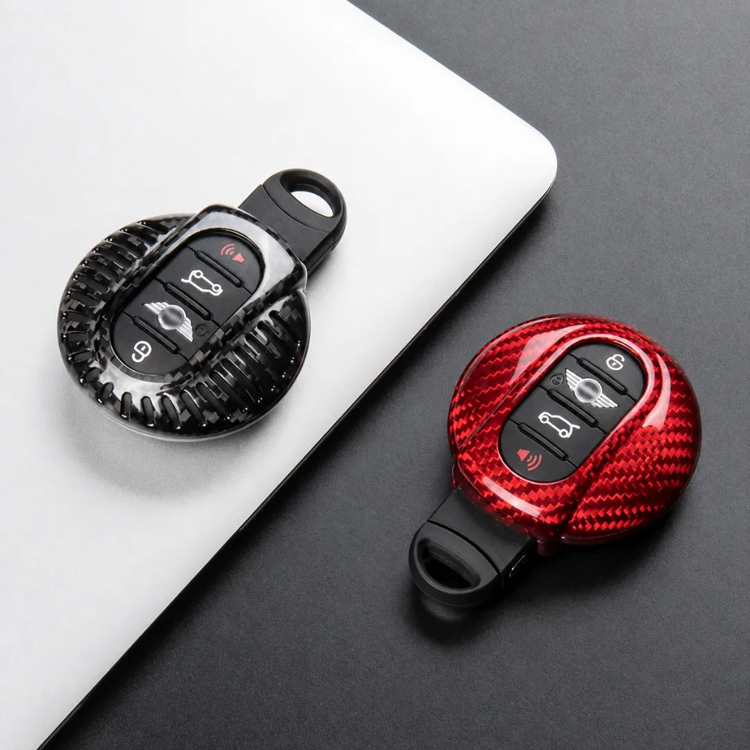 Two carbon-fiber-patterned keychain car key fobs shown on a dark surface, one resting on a white laptop lid and one alone. Both fobs have black button areas with vehicle control icons.