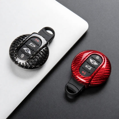 Two carbon-fiber-patterned keychain car key fobs shown on a dark surface, one resting on a white laptop lid and one alone. Both fobs have black button areas with vehicle control icons.
