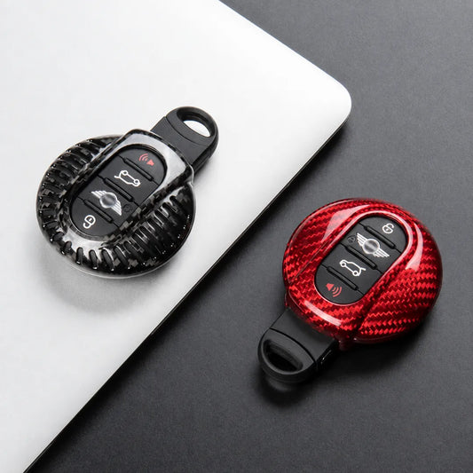 Two carbon-fiber-patterned keychain car key fobs shown on a dark surface, one resting on a white laptop lid and one alone. Both fobs have black button areas with vehicle control icons.