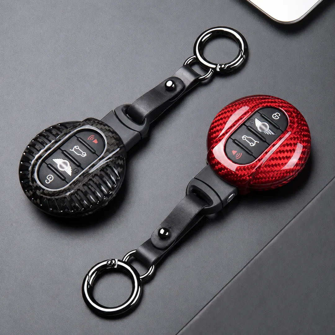 Two car key fobs with protective covers, one in black carbon fiber texture and one in red carbon fiber texture, both with black leather straps and silver key rings, displayed on a dark gray surface.