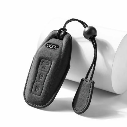 A black suede car key fob cover with a matching wristlet strap, shown on a white cylindrical prop against a white background. The cover features the embossed Audi four-ring logo.