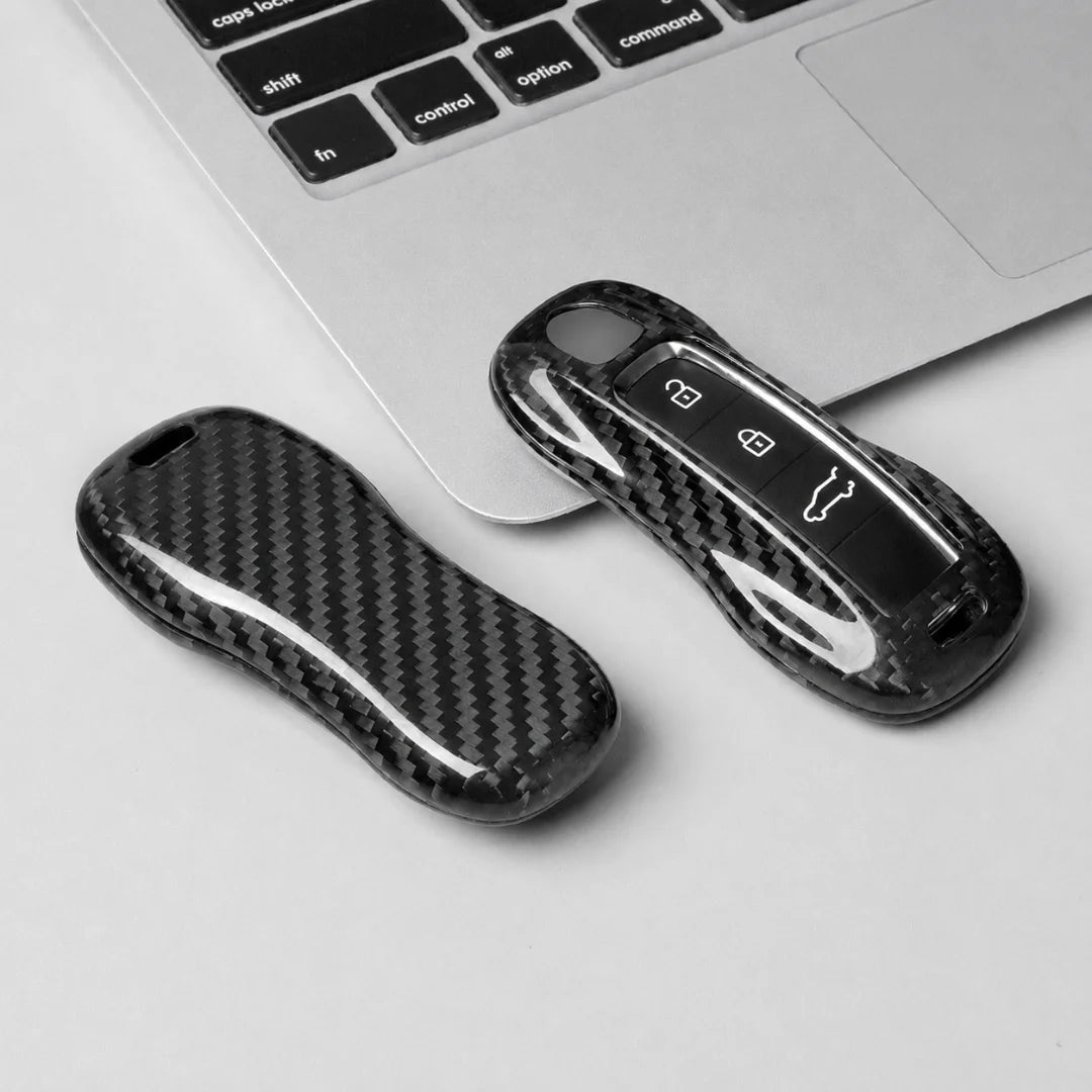 Two black carbon fiber keychain cases, one closed and one open showing the key fob inside, placed on a light gray surface next to a silver laptop keyboard.