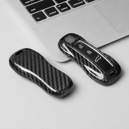 Two black carbon fiber keychain cases, one closed and one open showing the key fob inside, placed on a light gray surface next to a silver laptop keyboard.