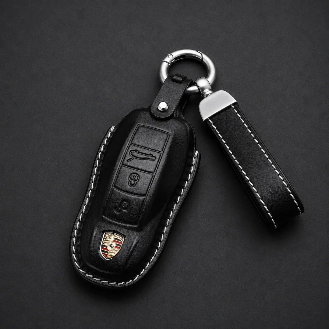 Black leather car key fob cover with white contrast stitching, a silver key ring, and a matching leather keychain strap, shown on a dark background. The cover features a small Porsche crest emblem near the bottom.