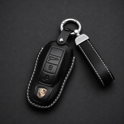 Black leather car key fob cover with white contrast stitching, a silver key ring, and a matching leather keychain strap, shown on a dark background. The cover features a small Porsche crest emblem near the bottom.