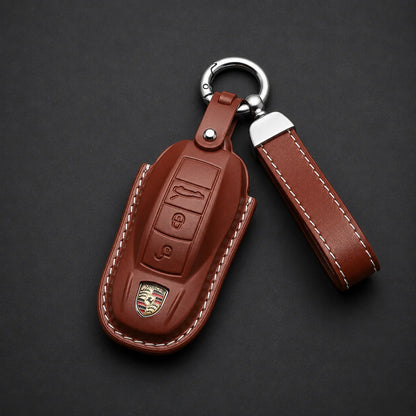 A brown leather car key fob cover with white contrast stitching, featuring an embossed Porsche crest and button details, attached to a silver key ring and matching leather keychain strap on a dark gray background.