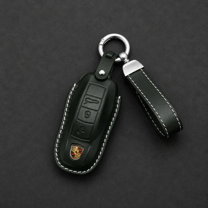 A dark forest green leather car key fob cover with white contrast stitching, featuring a silver metal key ring and a matching leather keychain loop. The cover is detailed with a metallic Porsche crest emblem near the bottom, set against a dark textured background.