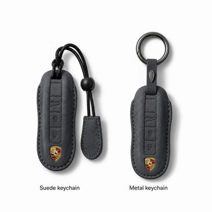 Two Porsche keychain covers shown on a light gray background, labeled 'Suede keychain' and 'Metal keychain.' The left version is suede-like dark gray with a black strap and a small Porsche crest near the bottom.