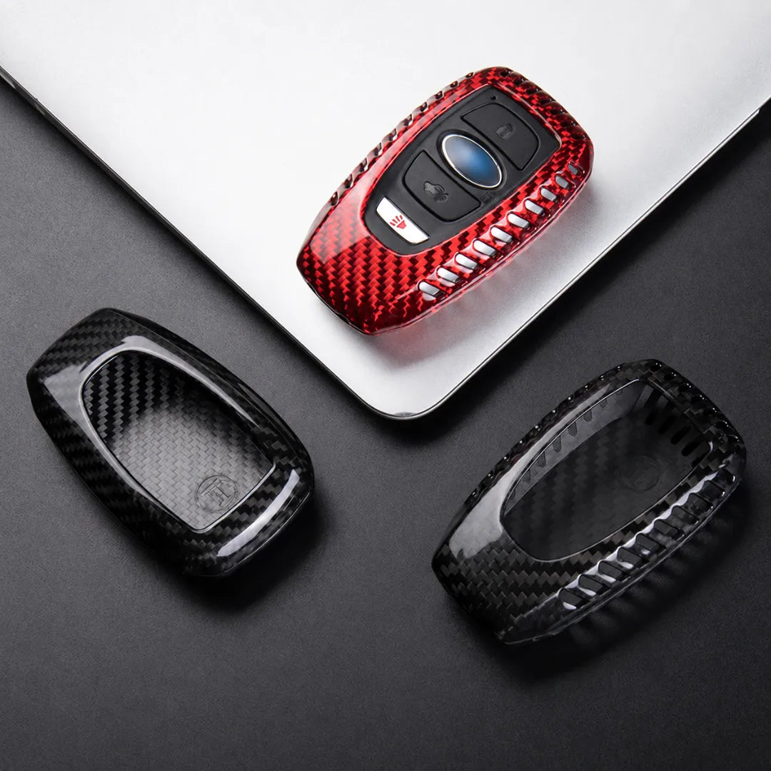 Three carbon fiber textured key fob covers in red, black, and clear are displayed on a dark surface with a silver laptop as a prop.