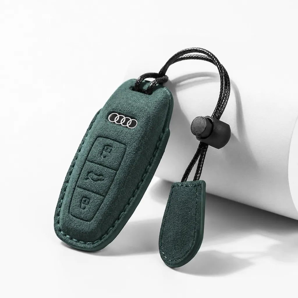 A dark green suede car key fob cover with an attached black cord and small tag, shown on a clean white background. The cover features an embossed silver Audi logo.