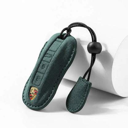 Dark green suede car key fob cover with a stitched edge, a small gold Porsche crest patch, and a black keychain cord with a black bead, shown on a white studio background.