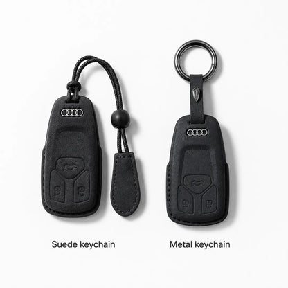 Two Audi keychain covers shown side by side on a light gray background: a suede keychain on the left with a black cord and tab, and a metal keychain on the right with a ring. Text below reads 'Suede keychain' and 'Metal keychain'.