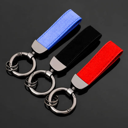 Three suede keychain fobs with silver metal key rings and swivel clips, featuring blue, black, and red fabric straps, arranged diagonally on a dark reflective surface.