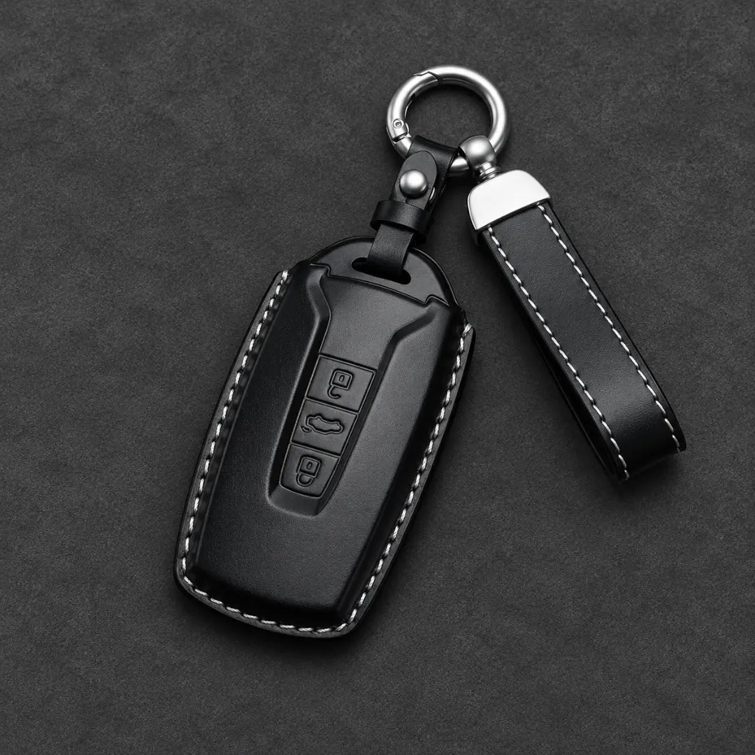Black leather car key fob cover with white contrast stitching, a silver key ring, and a matching leather keychain strap on a dark textured background.