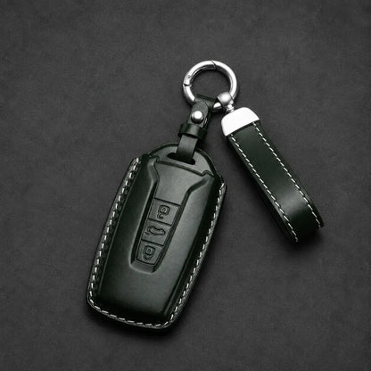 A dark green leather car key fob cover with white contrast stitching, shown with a matching leather keychain strap and silver metal key ring on a dark textured background.