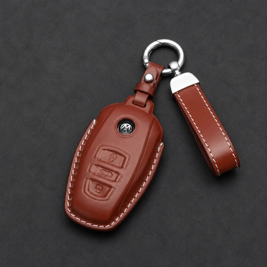 A brown leather car key fob cover with white contrast stitching, featuring an embossed logo on the front and a silver metal key ring and split ring, displayed on a dark textured background.