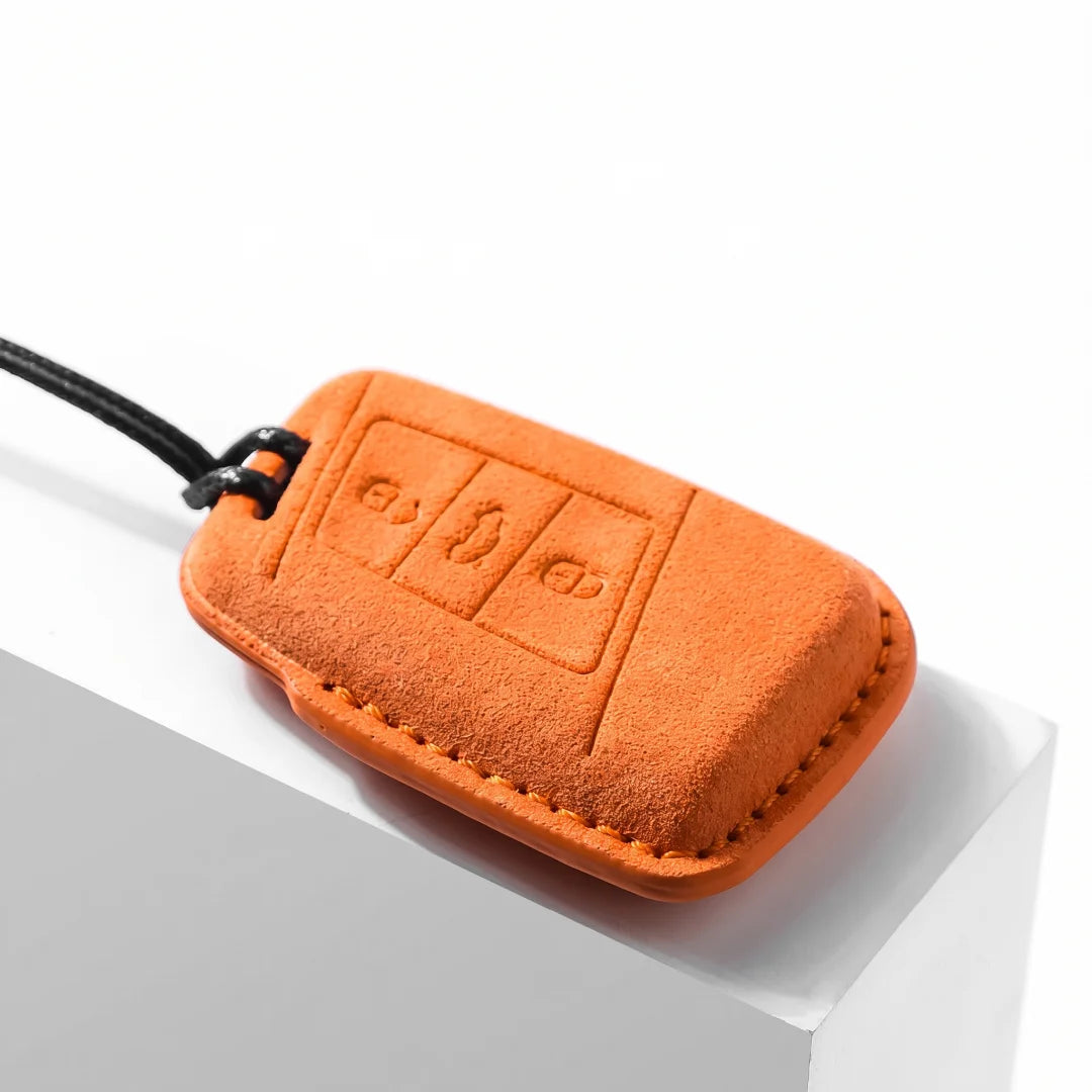 An orange suede car key fob cover with a black wrist strap, resting on a white geometric display block.