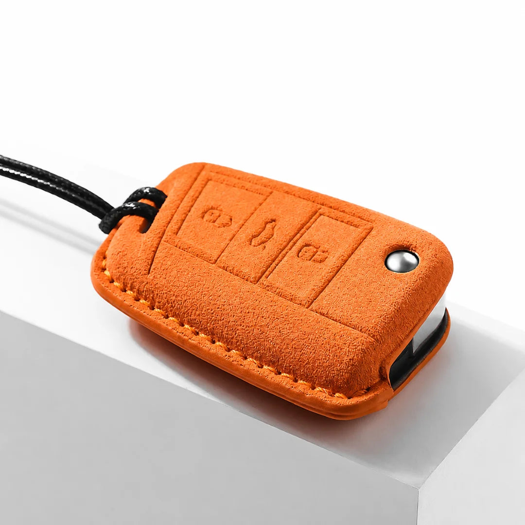 An orange suede car key fob cover with a black wrist strap, shown resting on a white geometric surface.